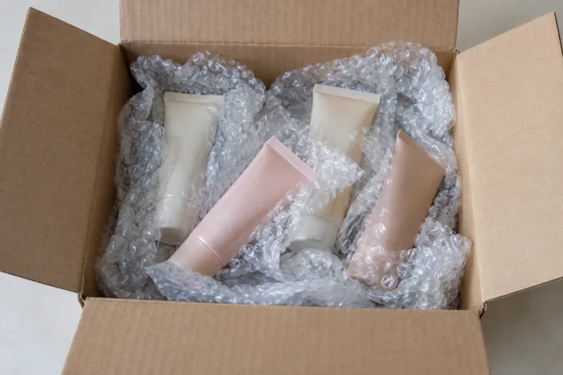Empat tube kosmetik berwarna krem dan merah muda dibungkus bubble wrap di dalam kardus terbuka